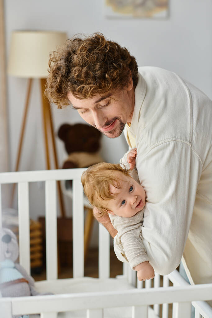 carinhoso pai encaracolado levantando seu filho bebê com olhos azuis do berço do bebê no berçário, paternidade - Foto, Imagem
