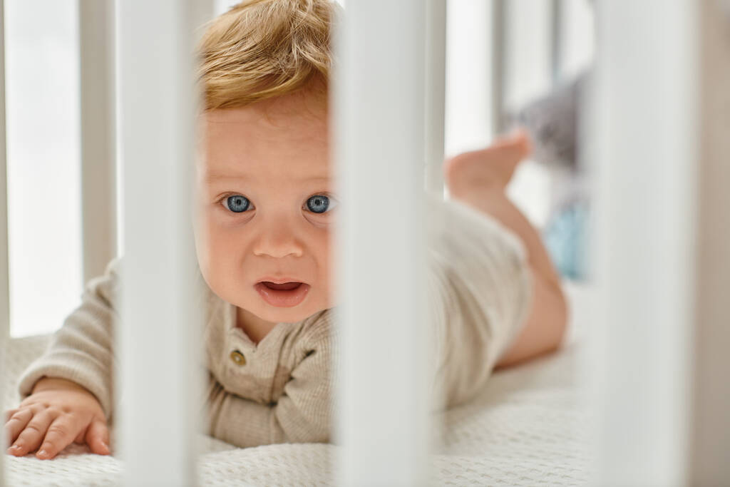 Bébé garçon aux yeux bleus regardant à travers les lamelles de lit dans sa chambre d'enfant, innocence de l'enfant - Photo, image