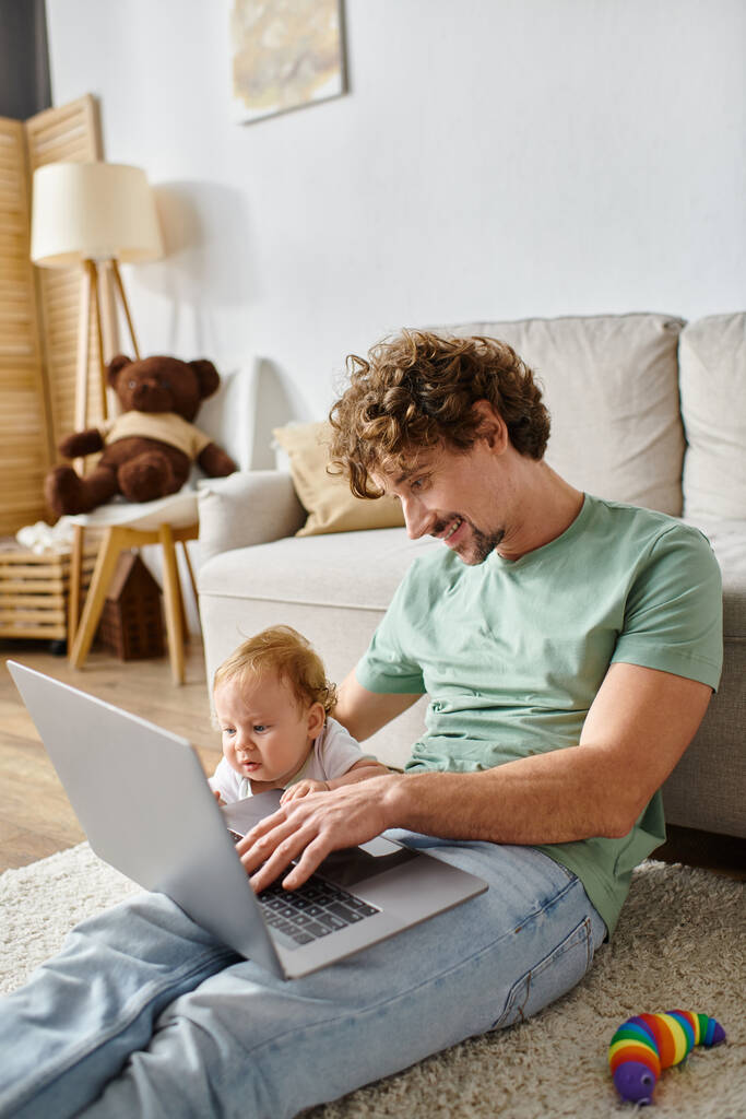 szczęśliwy ojciec za pomocą laptopa w pobliżu niemowlęcia syn w salonie, równoważąc między ojcostwem i pracy - Zdjęcie, obraz