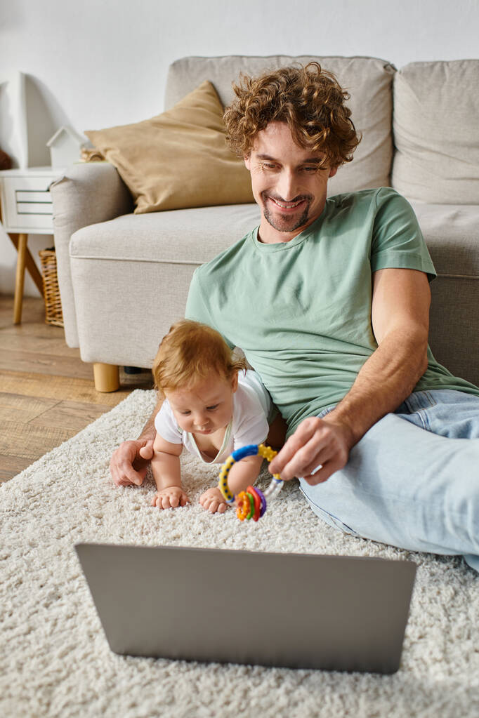 szczęśliwy człowiek trzymając grzechotka dziecko w pobliżu niemowlęcia syn i laptop na dywanie, równoważąc między pracą a życiem - Zdjęcie, obraz