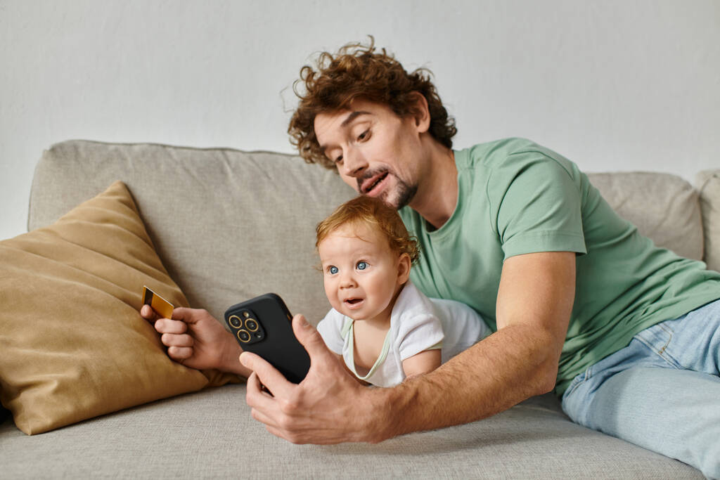smiling father shopping online with credit card and smartphone with infant baby son alongside him - Photo, Image