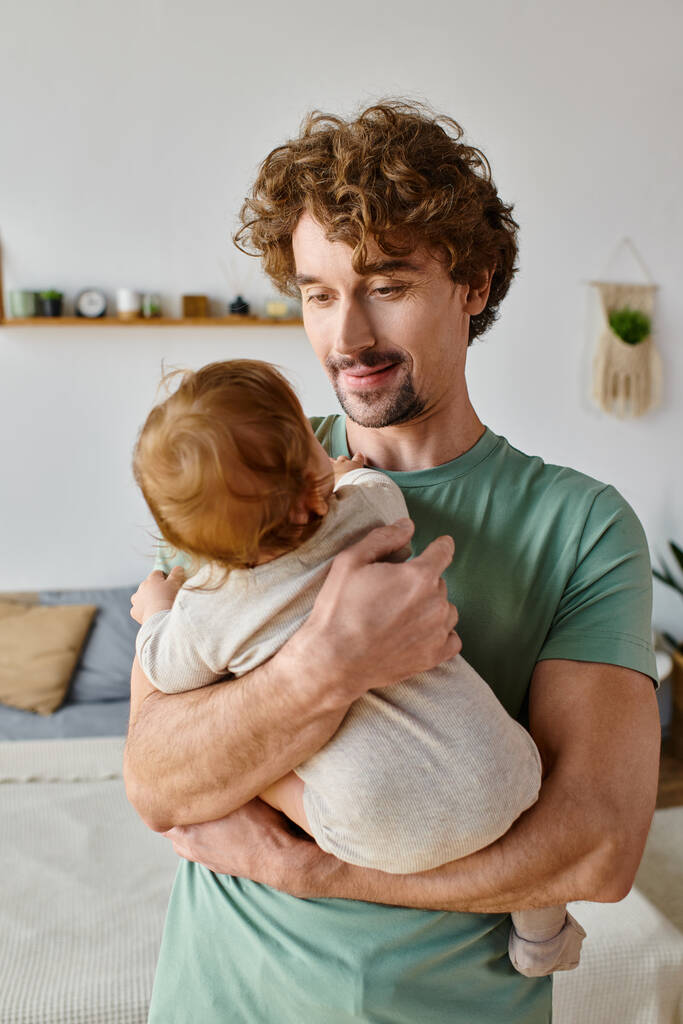 padre cariñoso con pelo rizado y barba sosteniendo en brazos a su niño con ropa de bebé, crianza - Foto, Imagen