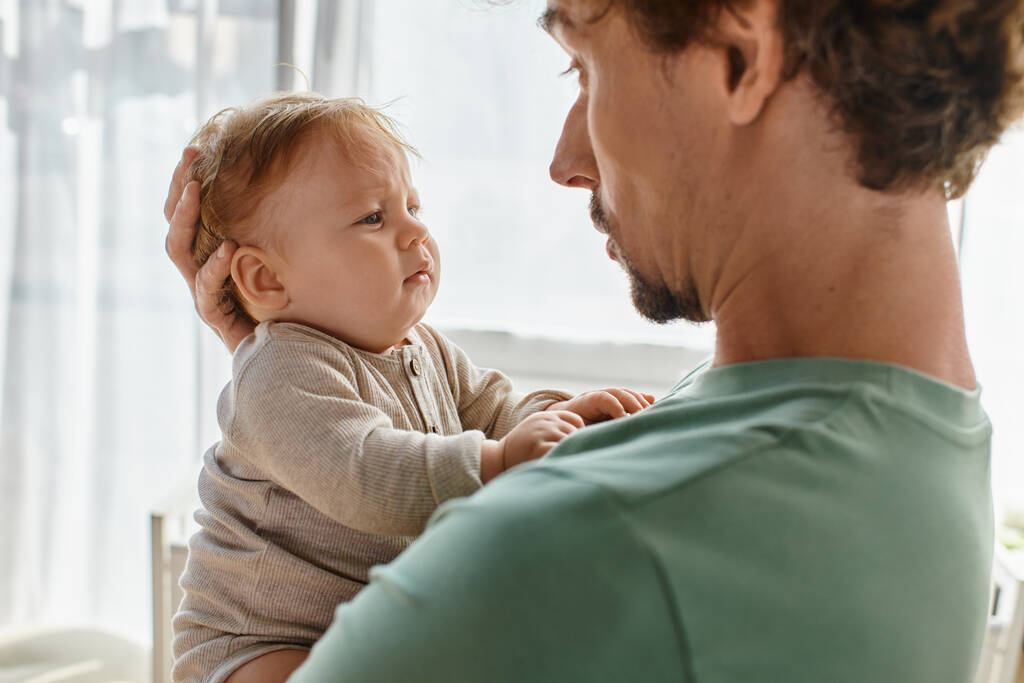 pai carinhoso com cabelo encaracolado e barba segurando nos braços seu menino em roupas de bebê, retrato - Foto, Imagem
