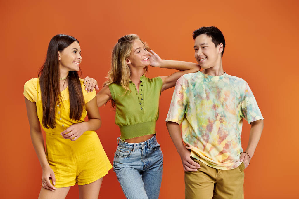 joyeux adolescent interracial amis dans lumineux tenues souriant à l 'autre dans orange toile de fond - Photo, image