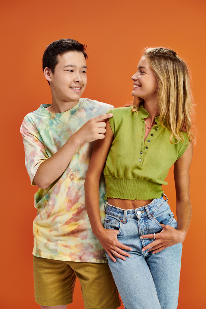 radosne międzyrasowe nastolatki w luźnych ubraniach uśmiechające się do siebie na pomarańczowym tle, przyjaźń - Zdjęcie, obraz