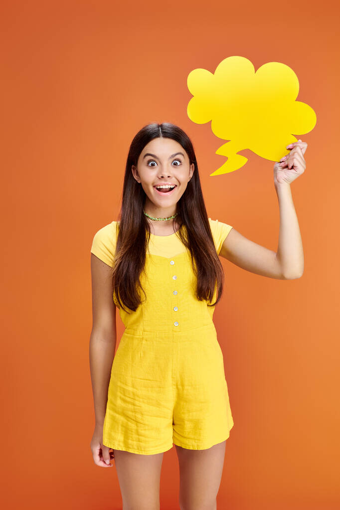 χαρούμενη έφηβη που ποζάρει με φούσκα σκέψης στο χέρι και κοιτάζει την κάμερα σε πορτοκαλί φόντο - Φωτογραφία, εικόνα
