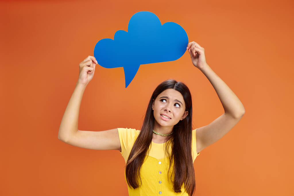 confused teenage girl in casual attire holding thought bubble above her head on orange backdrop - Photo, Image