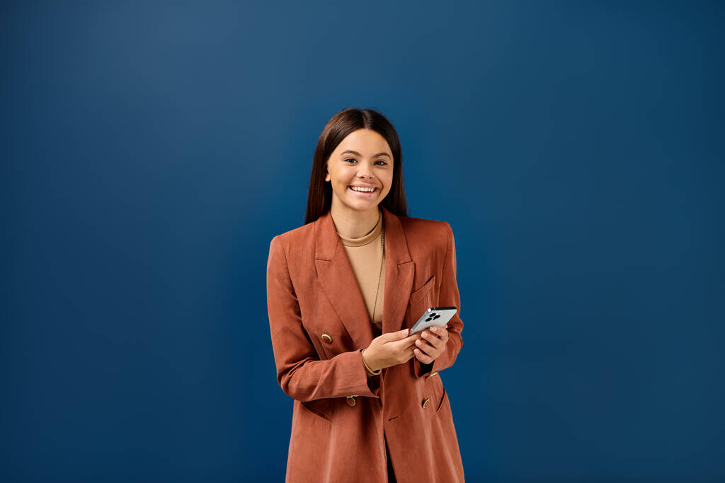 joyeuse adolescente à la mode en blazer brun tenant smartphone et souriant joyeusement à la caméra - Photo, image