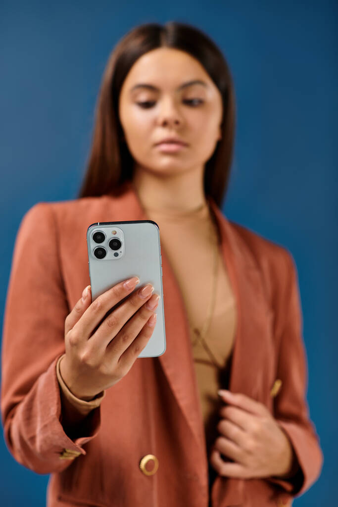 focus on smartphone in hands of stylish blurred teenage girl in brown blazer on dark blue backdrop - Photo, Image