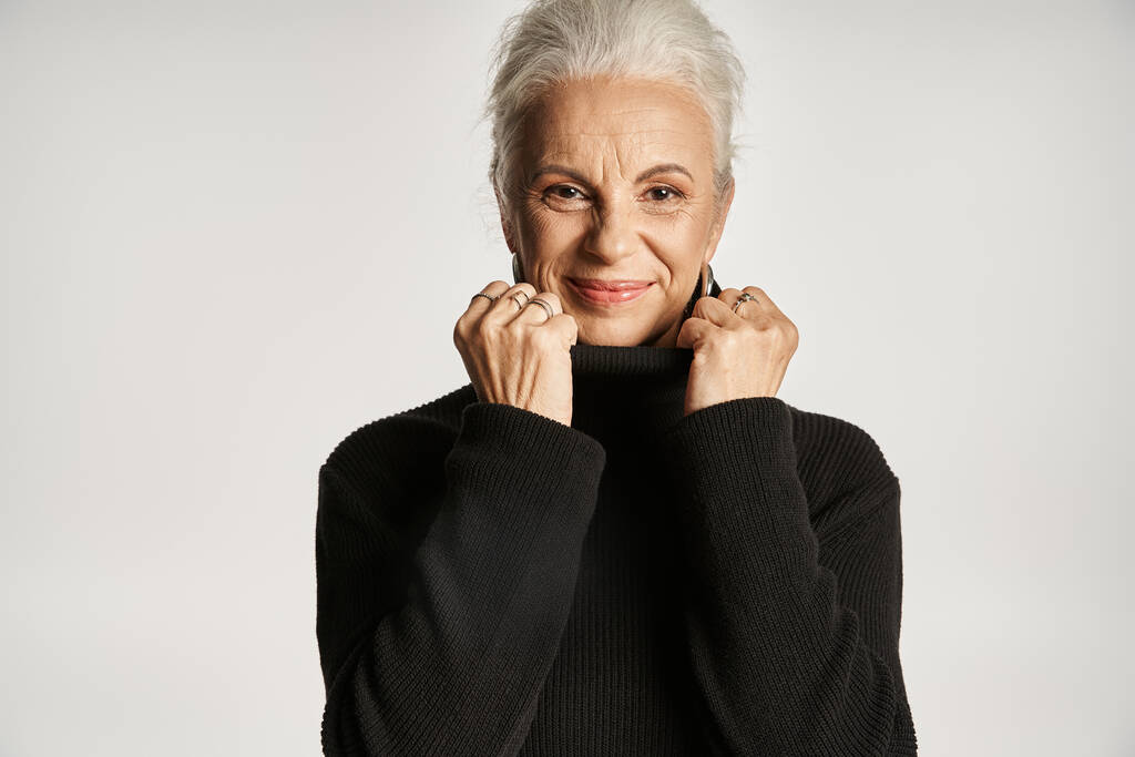 portrait of middle aged business woman adjusting collar of turtleneck sweater on grey backdrop - Photo, Image