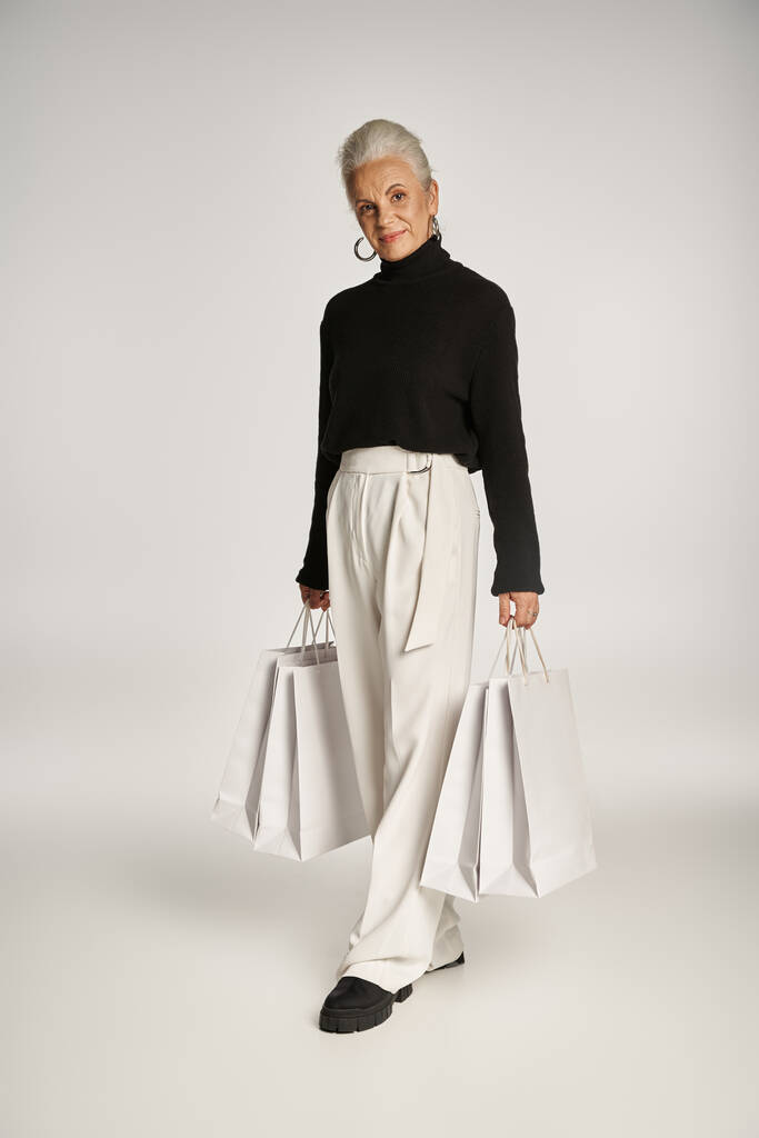 エレガントな服装とグレーのショッピングバッグで立っているフープイヤリングで幸せな中年女性 - 写真・画像