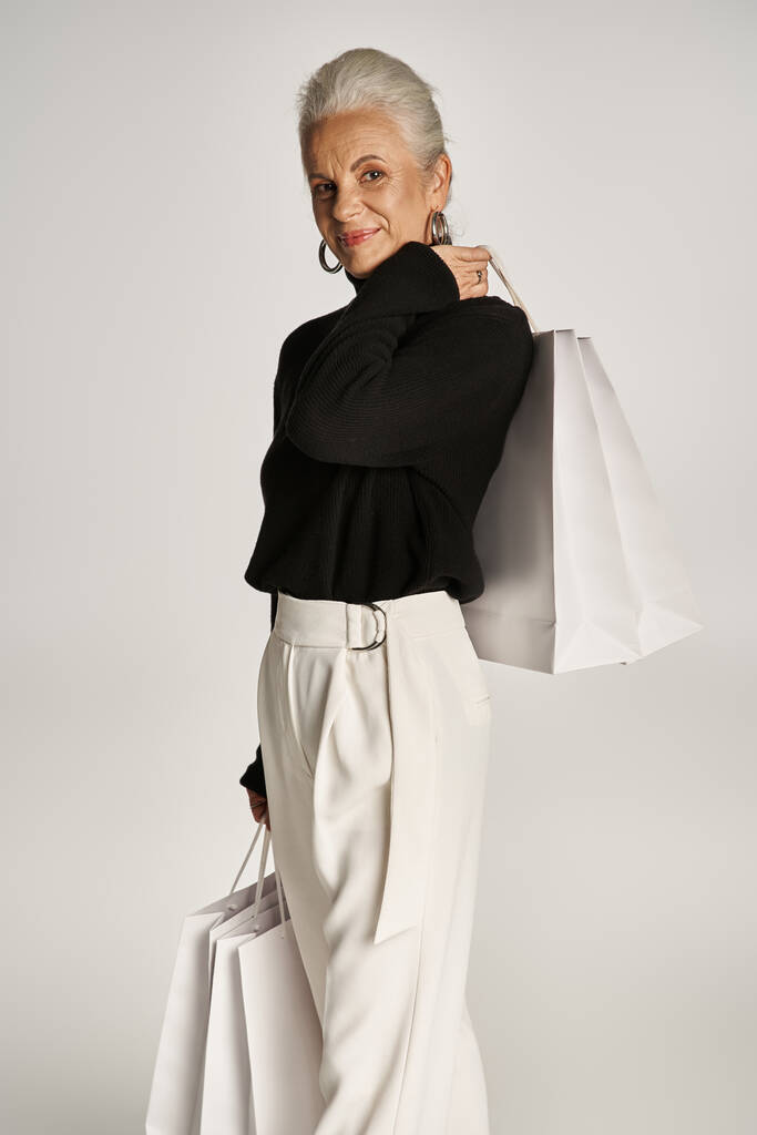 χαρούμενη μεσήλικη γυναίκα με κομψή ενδυμασία και σκουλαρίκια στεφάνι στέκεται με τσάντες για ψώνια σε γκρι - Φωτογραφία, εικόνα