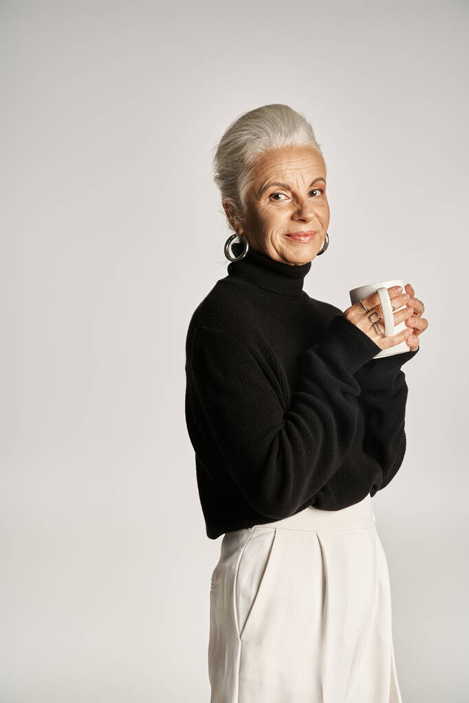 pleased middle aged woman in black turtleneck sweater holding cup of coffee on grey backdrop - Photo, Image