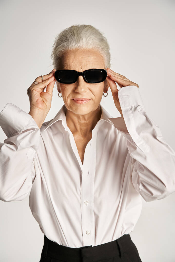 elegante donna di mezza età con i capelli grigi che indossa occhiali da sole e in piedi in elegante abbigliamento su grigio - Foto, immagini