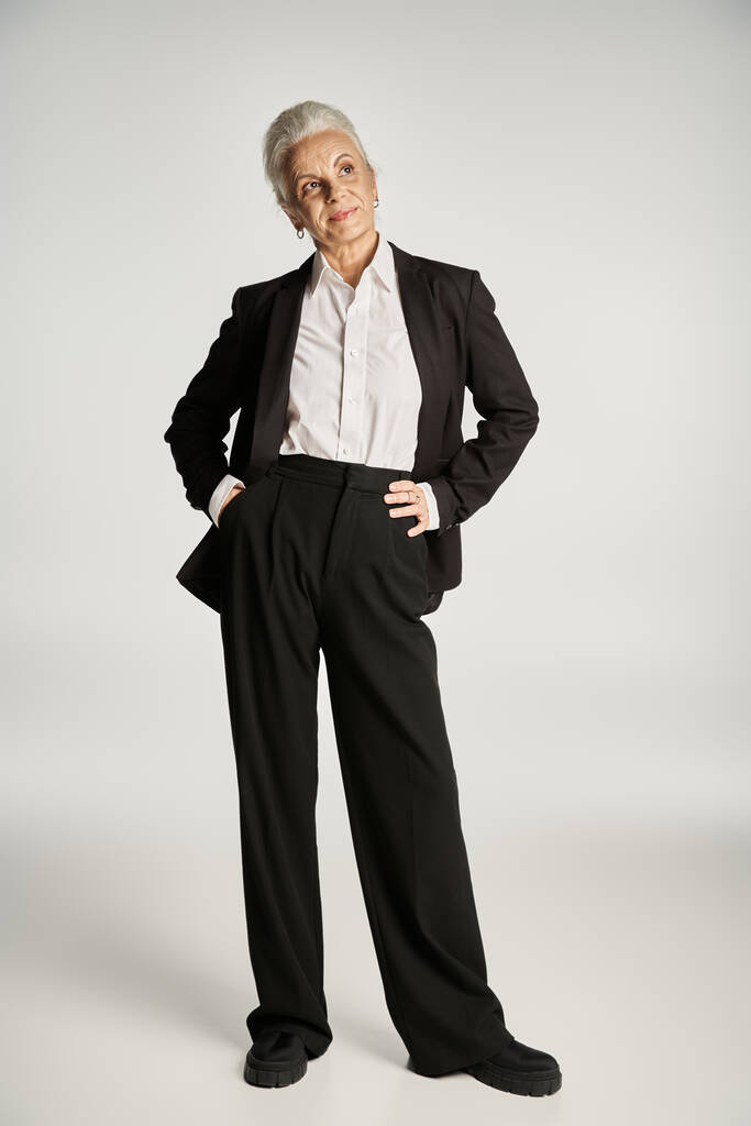 bela e de cabelos grisalhos mulher de meia idade em traje elegante posando em fundo cinza - Foto, Imagem