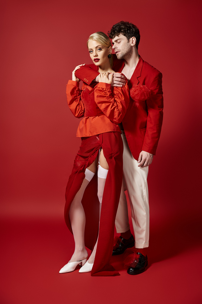 hombre guapo abrazando mujer rubia en traje elegante sobre fondo rojo, pareja de moda - Foto, Imagen