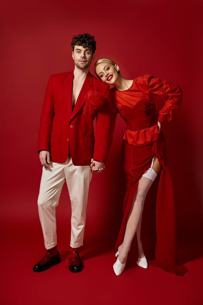 glückliche blonde Frau hält die Hände eines gutaussehenden Mannes in stilvoller Kleidung auf rotem Hintergrund, Mode - Foto, Bild