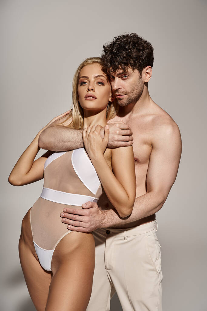 Мужчина без рубашки с мускулистым телом, обнимающий соблазнительную женщину на сером фоне, сексуальная пара - Фото, изображение