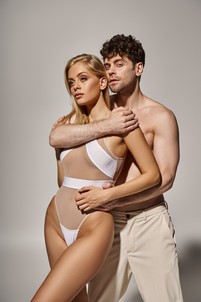 Мужчина без рубашки с мускулистым телом, обнимающий красивую молодую женщину на сером фоне, сексуальная пара - Фото, изображение