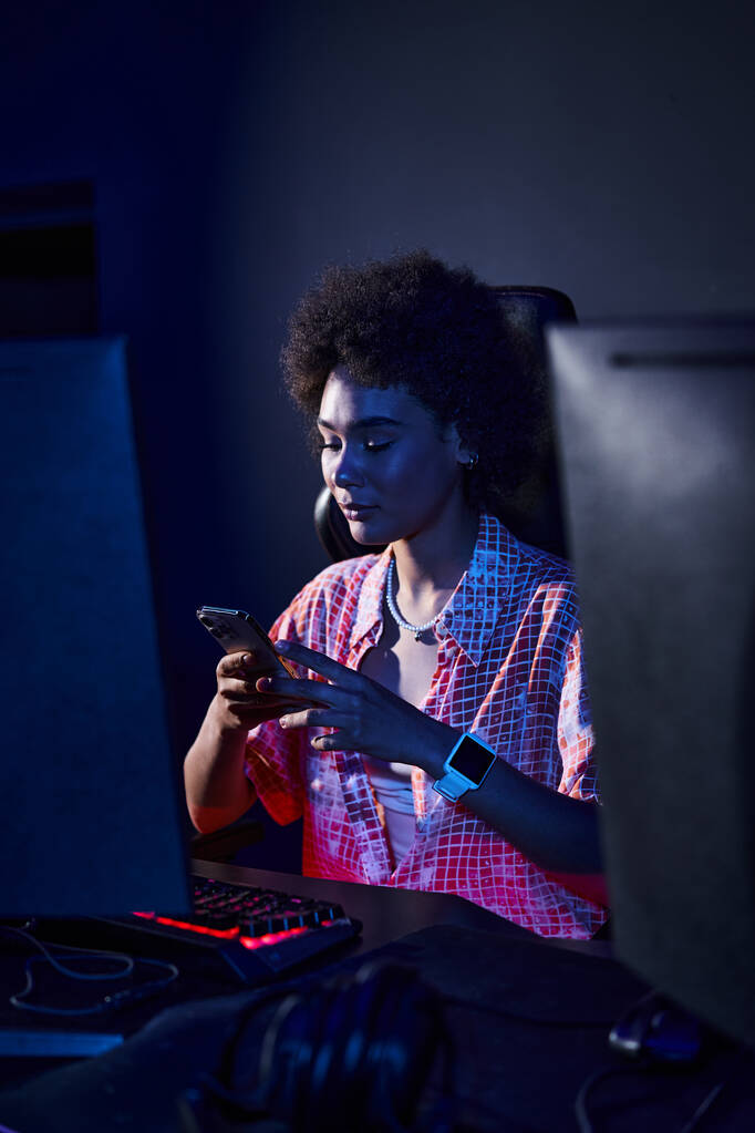 Bilgisayar monitöründen mavi ışık saçan kıvırcık saçlı Afro-Amerikan kadın akıllı telefonunu kullanıyor. - Fotoğraf, Görsel