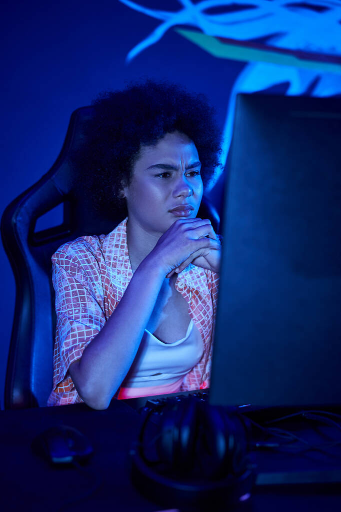 konzentriert afrikanisch-amerikanische Frau spielt intensiv in einem blau beleuchteten Raum, Cybersport-Konzept - Foto, Bild