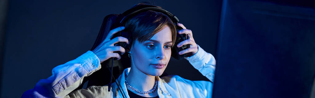 fokussierte Frau mit Kopfhörern und Blick auf den Computer in einem blau beleuchteten Raum, Cybersport-Spiel Banner - Foto, Bild