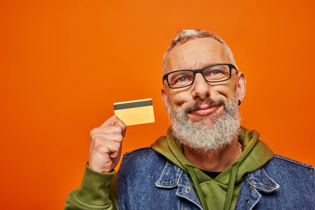 alegre buen aspecto hombre maduro en ropa vibrante con gafas celebración de la tarjeta de crédito cerca de su cara - Foto, Imagen