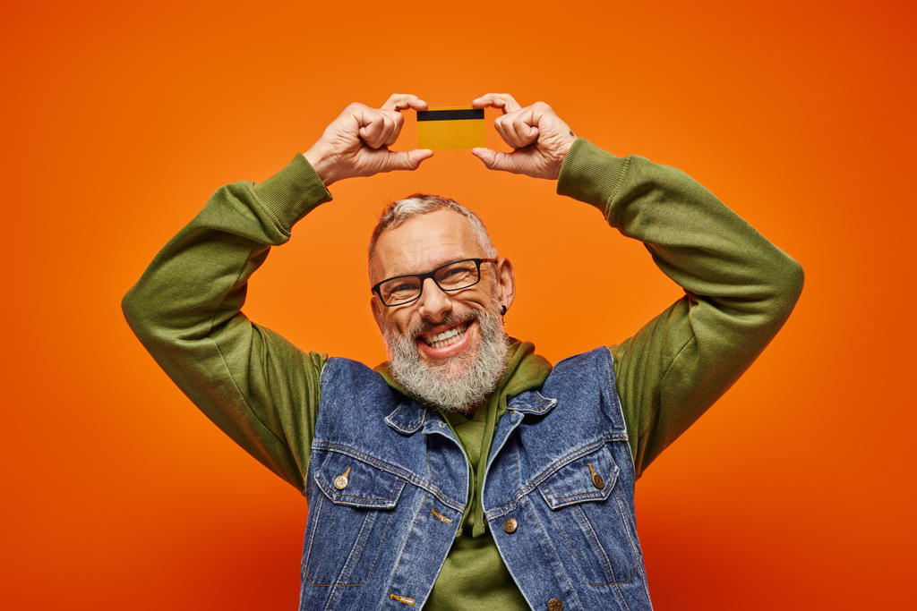 vrolijke volwassen man in levendige groene hoodie houden credit card boven het hoofd en glimlachen op de camera - Foto, afbeelding