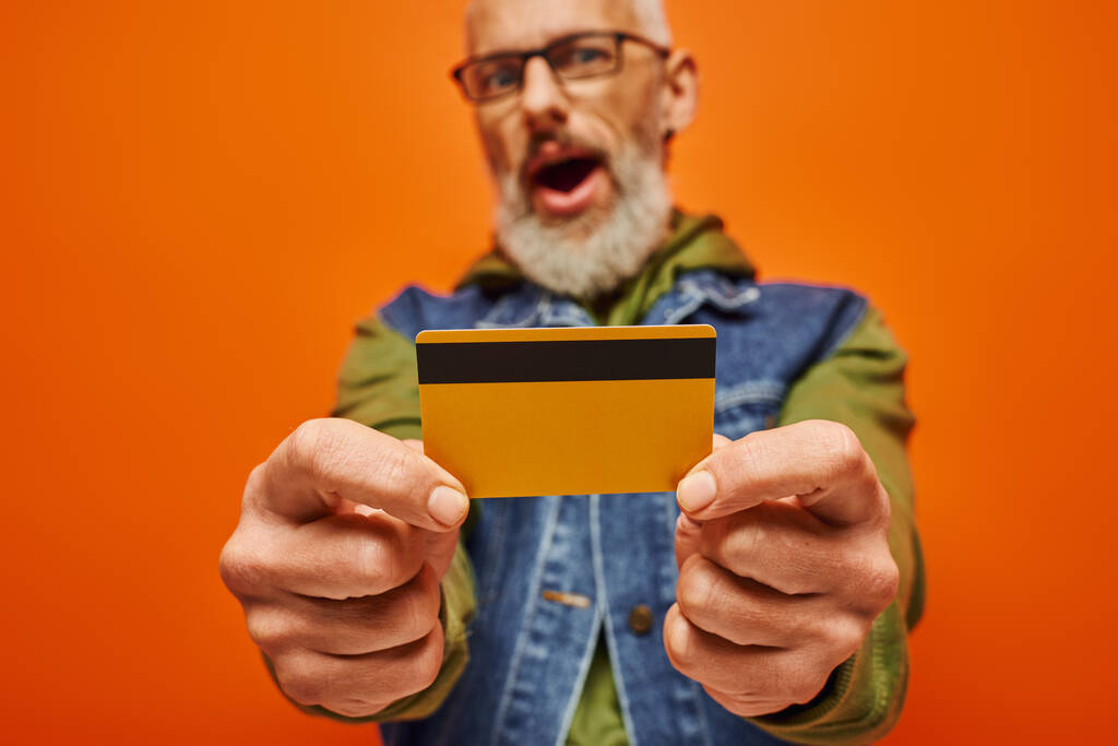 zaměření na kreditní kartu v rukou zralého rozmazané vousatý muž v živé oblečení na oranžovém pozadí - Fotografie, Obrázek