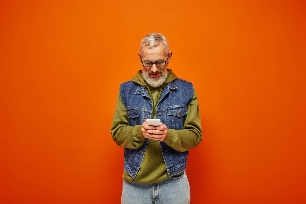 przystojny funky Dojrzałe mężczyzna w zielony modny bluza patrząc na smartfon na pomarańczowym tle - Zdjęcie, obraz