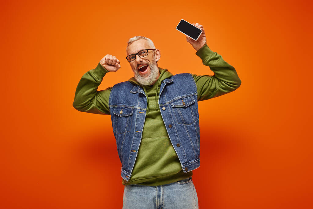 vrolijke aantrekkelijke volwassen man in groene hoodie en denim vest holding telefoon en kijken naar camera - Foto, afbeelding