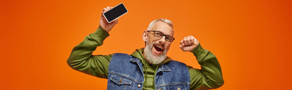 Весело красивый зрелый мужчина в зеленой толстовке и джинсовой жилетке держит телефон и смотрит в камеру, баннер - Фото, изображение