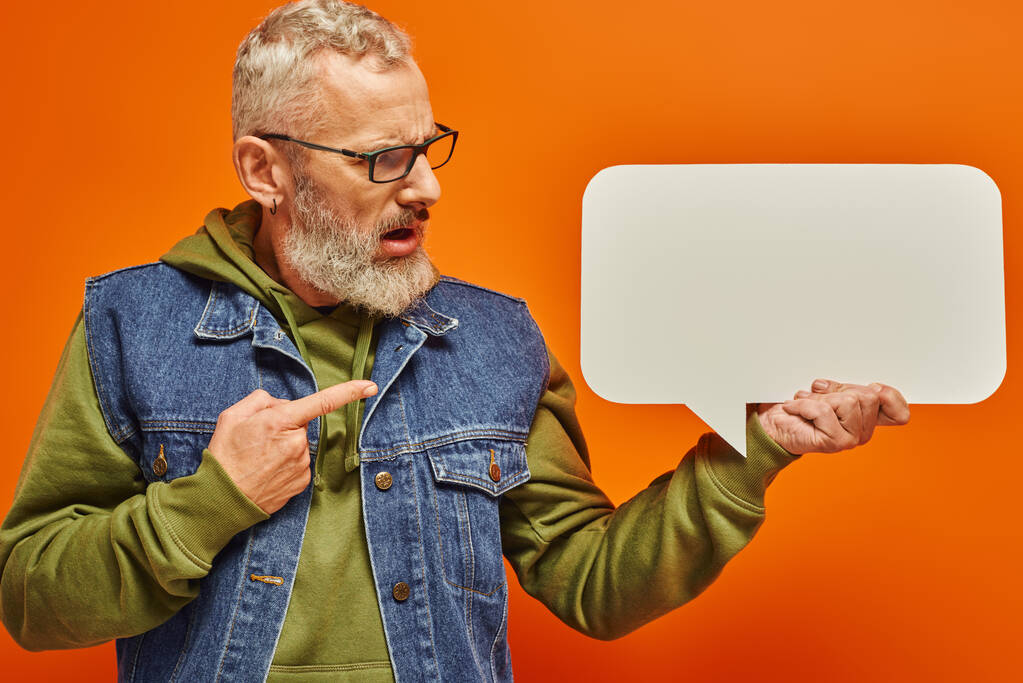 Verwirrter gutaussehender älterer Mann in Jeansweste blickt auf Sprechblase in der Hand vor orangefarbenem Hintergrund - Foto, Bild