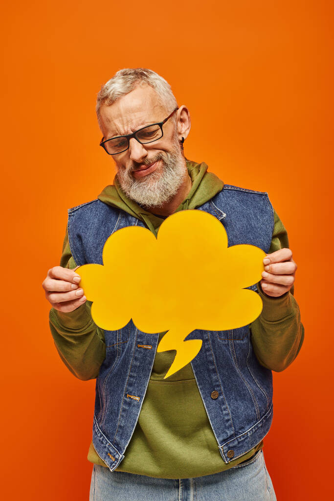 confused upset mature man in denim vest looking at thought bubble in his hands on orange backdrop - Photo, Image
