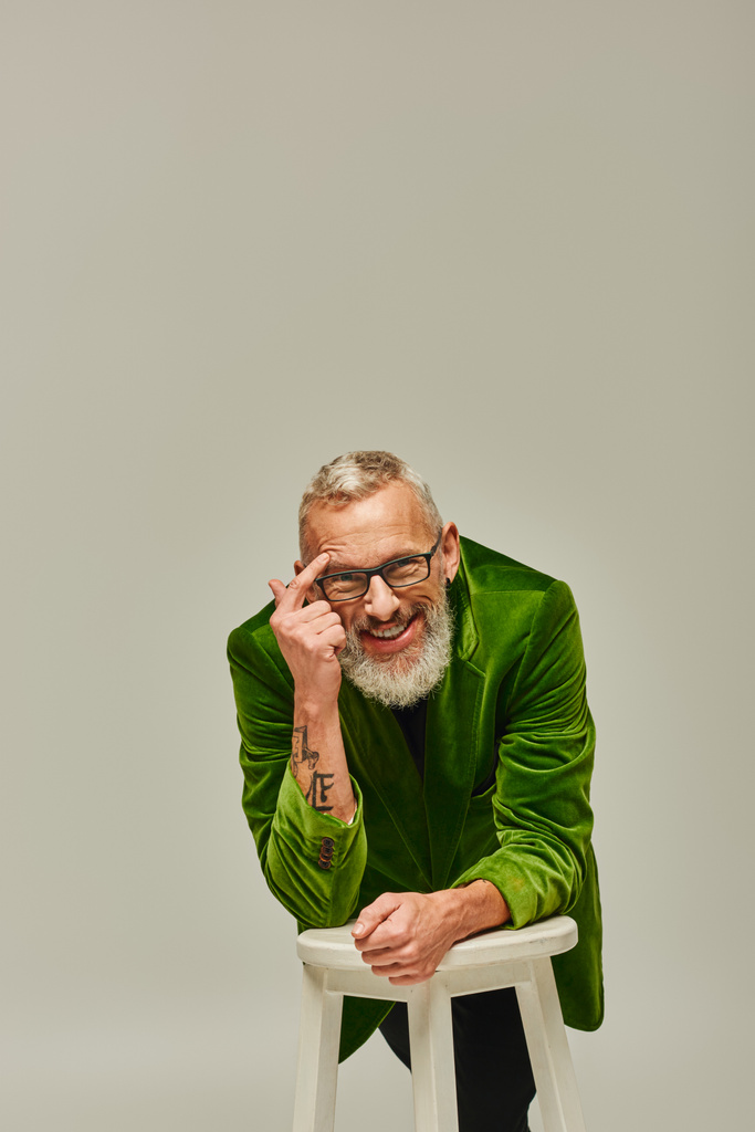 joyeux barbu mature homme en tenue vive avec des tatouages posant avec chaise haute et souriant à la caméra - Photo, image