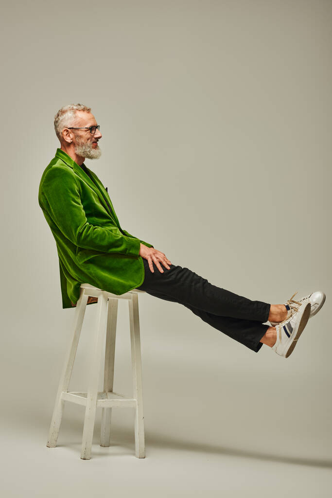 knap funky volwassen man model in heldere kledij met accessoires poseren op hoge stoel in profiel - Foto, afbeelding