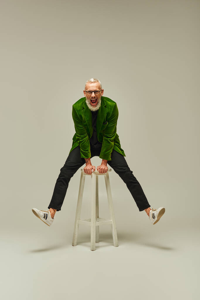 joyful dobře vypadající dospělý muž v pulzující oblečení pózovat na vysoké židli aktivně a usmívat se na kameru - Fotografie, Obrázek