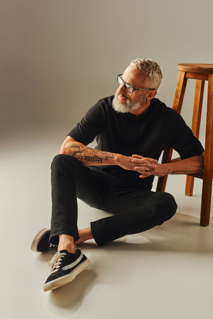 ελκυστικός ώριμος άντρας σε μαύρο t-shirt με γυαλιά και τατουάζ κάθεται στο πάτωμα δίπλα σε ψηλή καρέκλα - Φωτογραφία, εικόνα