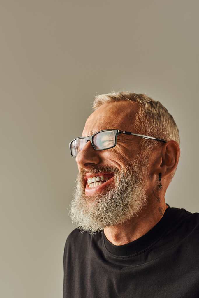 Porträt eines fröhlichen reifen Mannes mit Tätowierung in schwarzem T-Shirt, der glücklich auf beigem Hintergrund lächelt - Foto, Bild