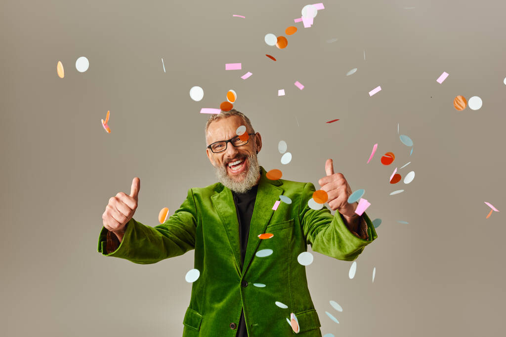 joyful starší muž model v zeleném blejzru ukazující palce nahoru pod konfety déšť na béžové pozadí - Fotografie, Obrázek