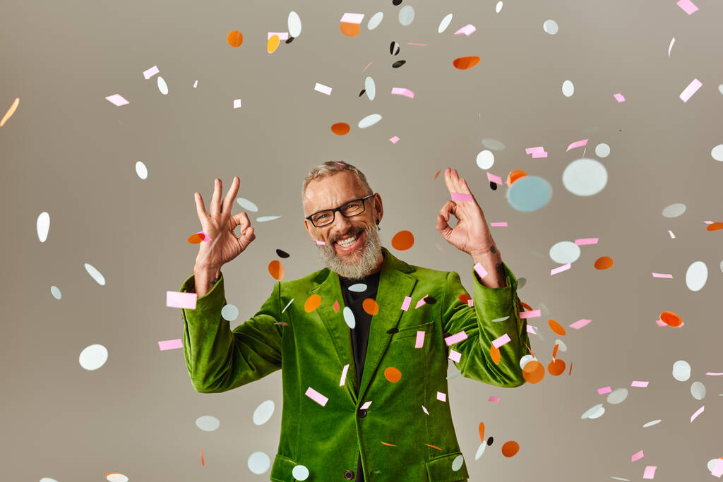 mooie vrolijke volwassen man in groene blazer tonen oke teken onder confetti regen op beige achtergrond - Foto, afbeelding