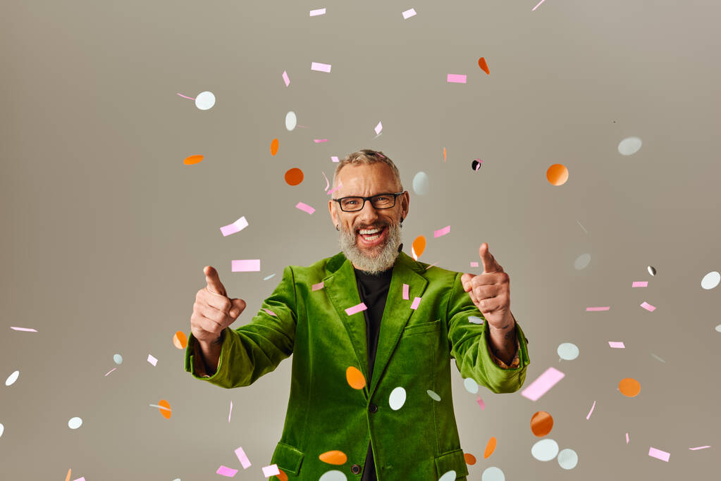 joyous handsome mature man in green blazer pointing at camera under confetti rain on beige backdrop - Photo, Image