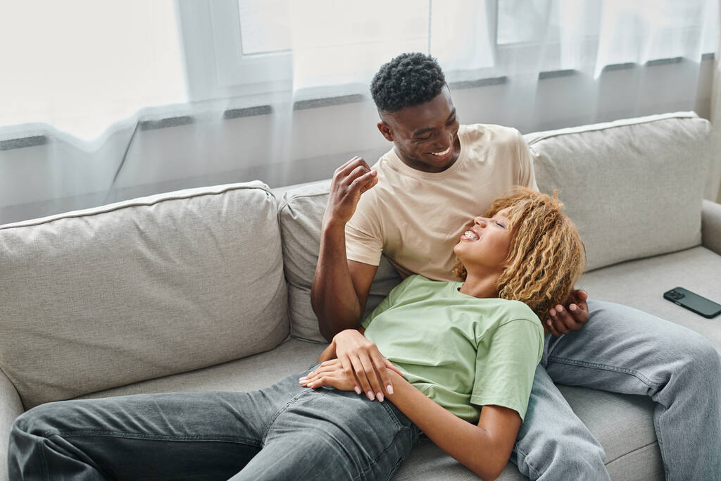 gelukkig Afrikaans Amerikaans paar samen lachen terwijl ontspannen op de bank in de woonkamer, luchthartig - Foto, afbeelding