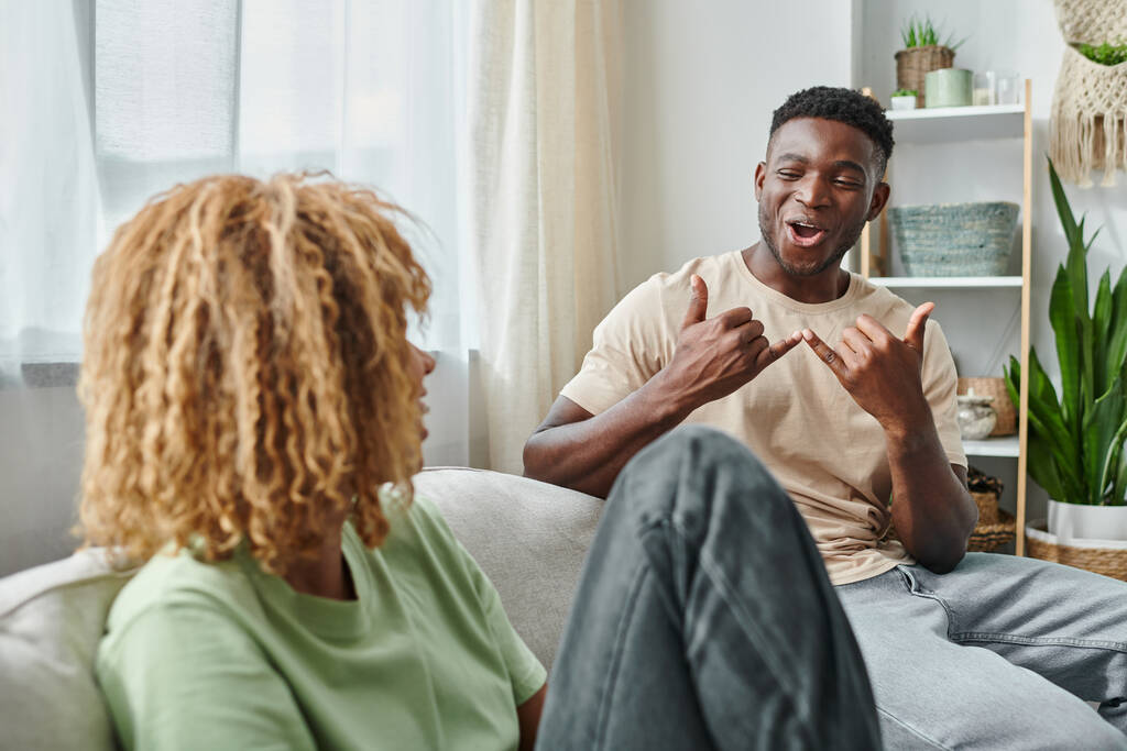 freudiger schwarzer Mann lächelt, während er mit Freundin im Wohnzimmer mit Gebärdensprache kommuniziert - Foto, Bild