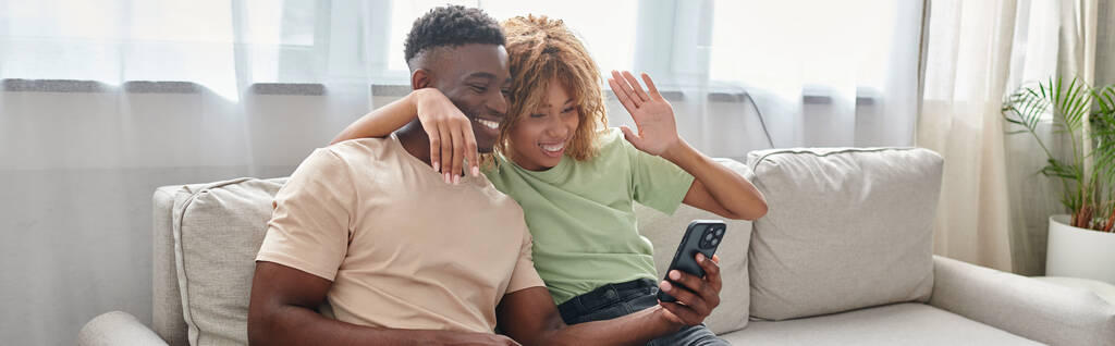 šťastný africký americký pár video chatování na smartphone zatímco sedí na gauči spolu, banner - Fotografie, Obrázek