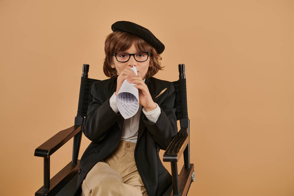 κομψό αγόρι σε μπερέ και γυαλιά κάθεται στην καρέκλα διευθυντής, μιλώντας σε κομμάτι χαρτί έλασης - Φωτογραφία, εικόνα