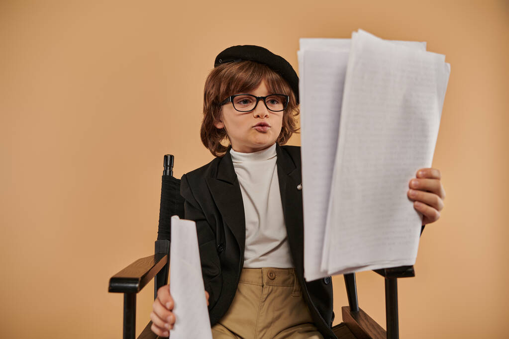 メガネの忙しい少年は,ディレクターの椅子に座り,新聞を見て,スタジオで彼の読書に吸収される - 写真・画像