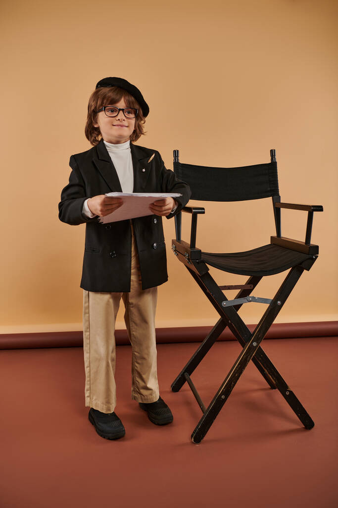 χαρούμενο αγόρι στέκεται περήφανα δίπλα στην καρέκλα του σκηνοθέτη, ντυμένο με σακάκι και παντελόνι, κρατώντας χαρτιά - Φωτογραφία, εικόνα