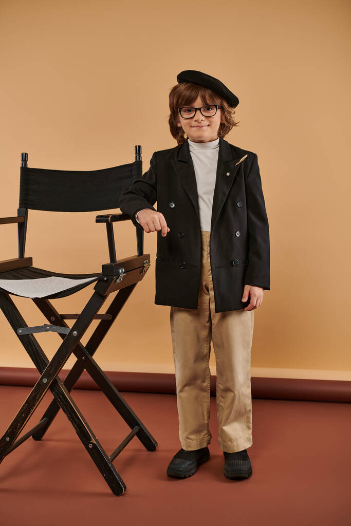 niño feliz se para orgullosamente al lado de la silla del director, usando abrigo y pantalones, con una gran sonrisa en la cara - Foto, imagen
