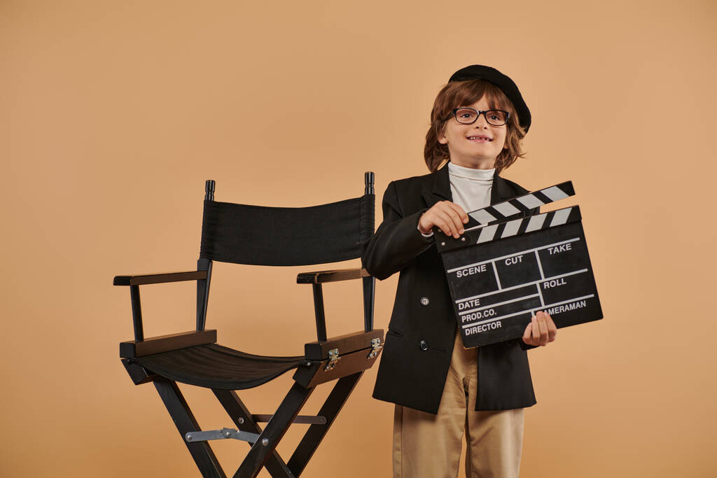 νεαρός κινηματογραφιστής με μοντέρνα ρούχα, ποζάρει χαρούμενος με μια σανίδα στο χέρι ενάντια σε μπεζ τοίχο - Φωτογραφία, εικόνα